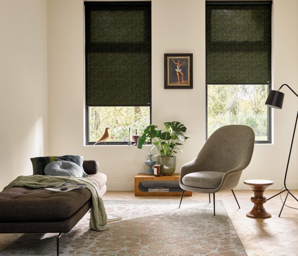 Woonkamer in natuurlijke stijl met vloerkleed met print, bankje en leesstoel, licht transparante rolgordijnen in donkergroen voor de hoge ramen. Woonkamer in natuurlijke stijl met vloerkleed met print, bankje en leesstoel, licht transparante rolgordijnen in donkergroen voor de hoge ramen.