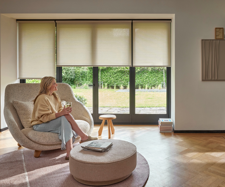 Beige/licht gele rolgordijnen in de woonkamer met een dame op een stoel Beige/licht gele rolgordijnen in de woonkamer met een dame op een stoel