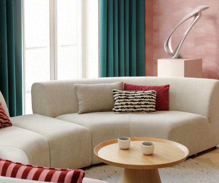 Blauw/groene gordijnen, roze behang en ronde grijze bank in woonkamer