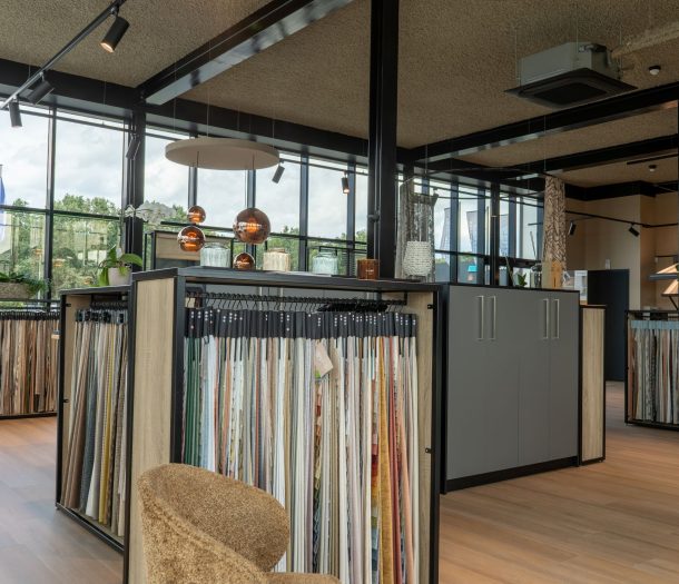Rekken vol gordijnstalen in alle kleuren bij Althoff Woonmode, met koperen hanglampen en grote ramen op de achtergrond