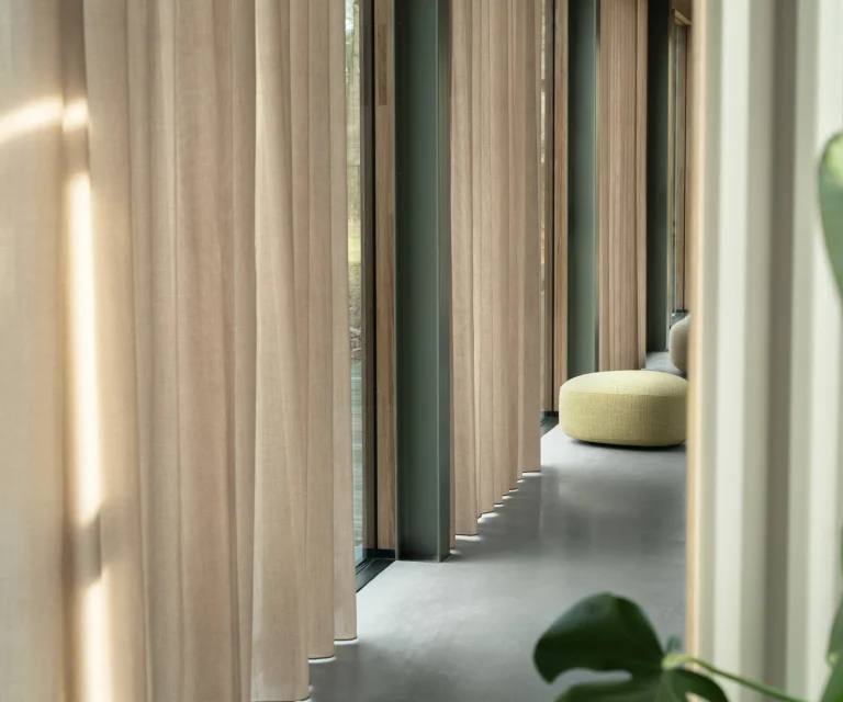 Beige Artex gordijnen met wave-plooi voor hoge ramen in een lichte gang met betonvloer