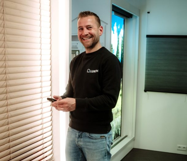 Lachende medewerker van Luken Wonen en Slapen met een telefoon in de hand naast een houten jaloezie in de showroom