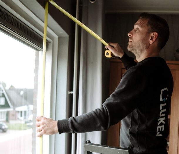 Medewerker van Luken Wonen en Slapen meet op een trapje een raam in met een rolmaat voor nieuwe raamdecoratie
