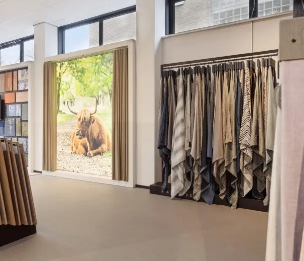 Showroom in Ede met een rek vol gordijnstalen in naturel en grijstinten, gordijnen voor een raam met een foto van een Schotse hooglander en vloerstalen