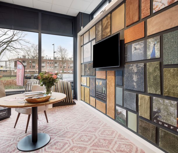 Tapijtwand met stalen in verschillende kleuren en structuren, met COREtec vloerstalen en een ronde tafel bij het raam van de showroom in Ede