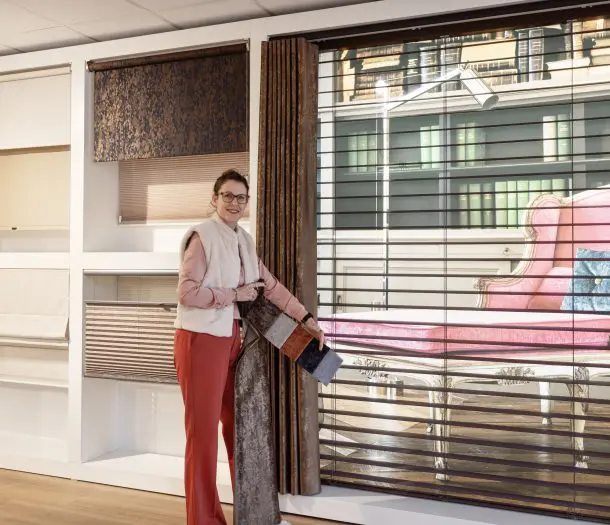 Medewerker laat gordijnstoffen zien in de showroom in Ede, naast een wand met raamdecoratie zoals houten jaloezieën, plissés en rolgordijnen