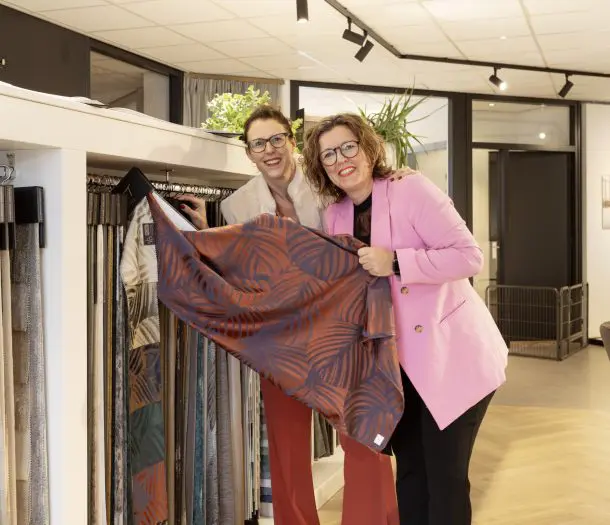 Twee medewerkers houden lachend een gordijnstof met een groot bladerprint in rood en blauw omhoog bij de stalenrekken